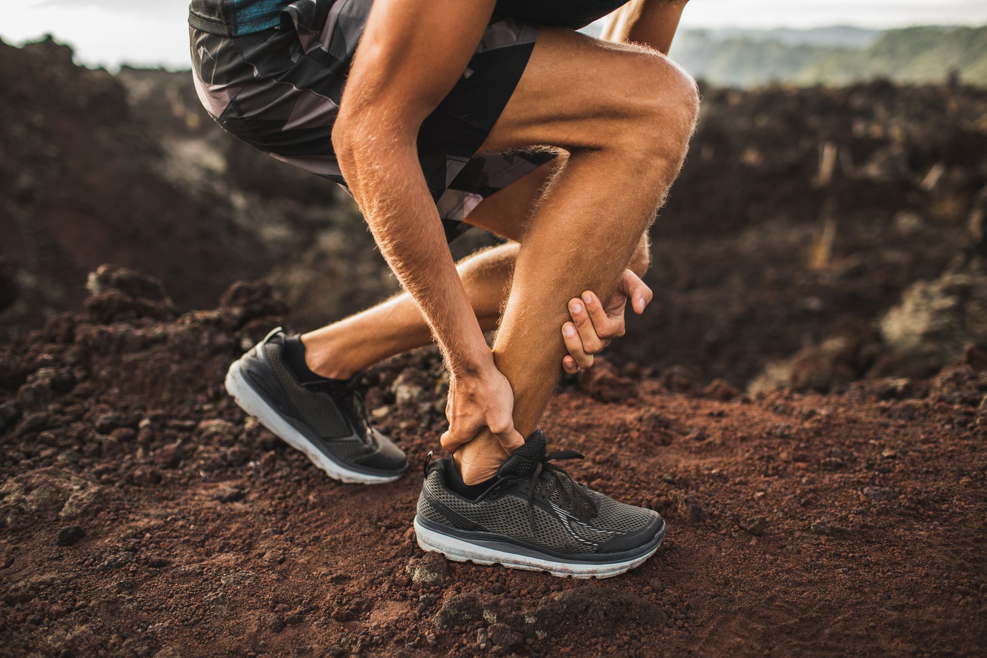 Person clutching ankle, likely injured, on a rocky trail wearing athletic shoes and black shorts.