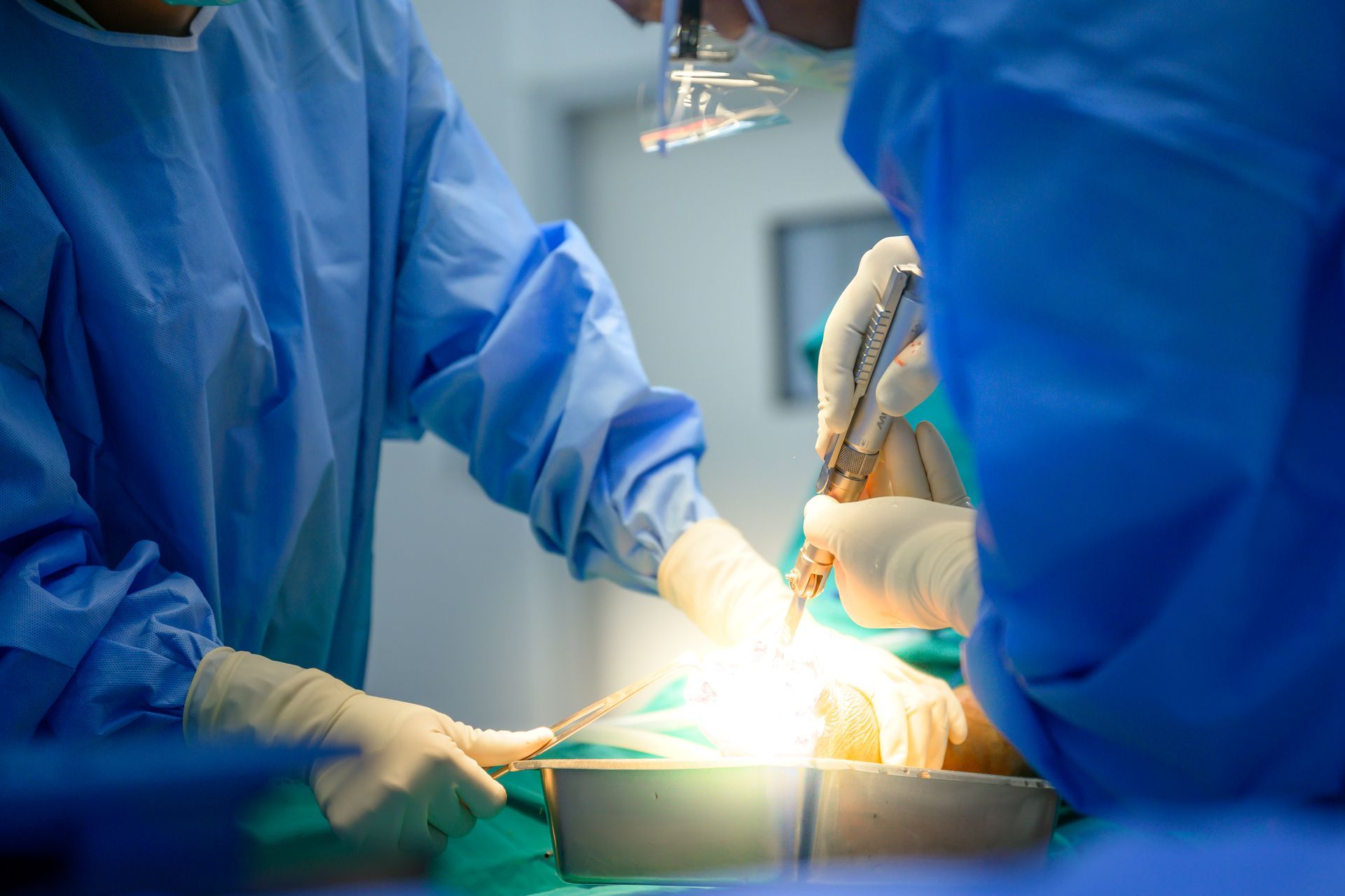 Surgeons in blue scrubs perform surgery in a brightly lit operating room, using medical instruments.