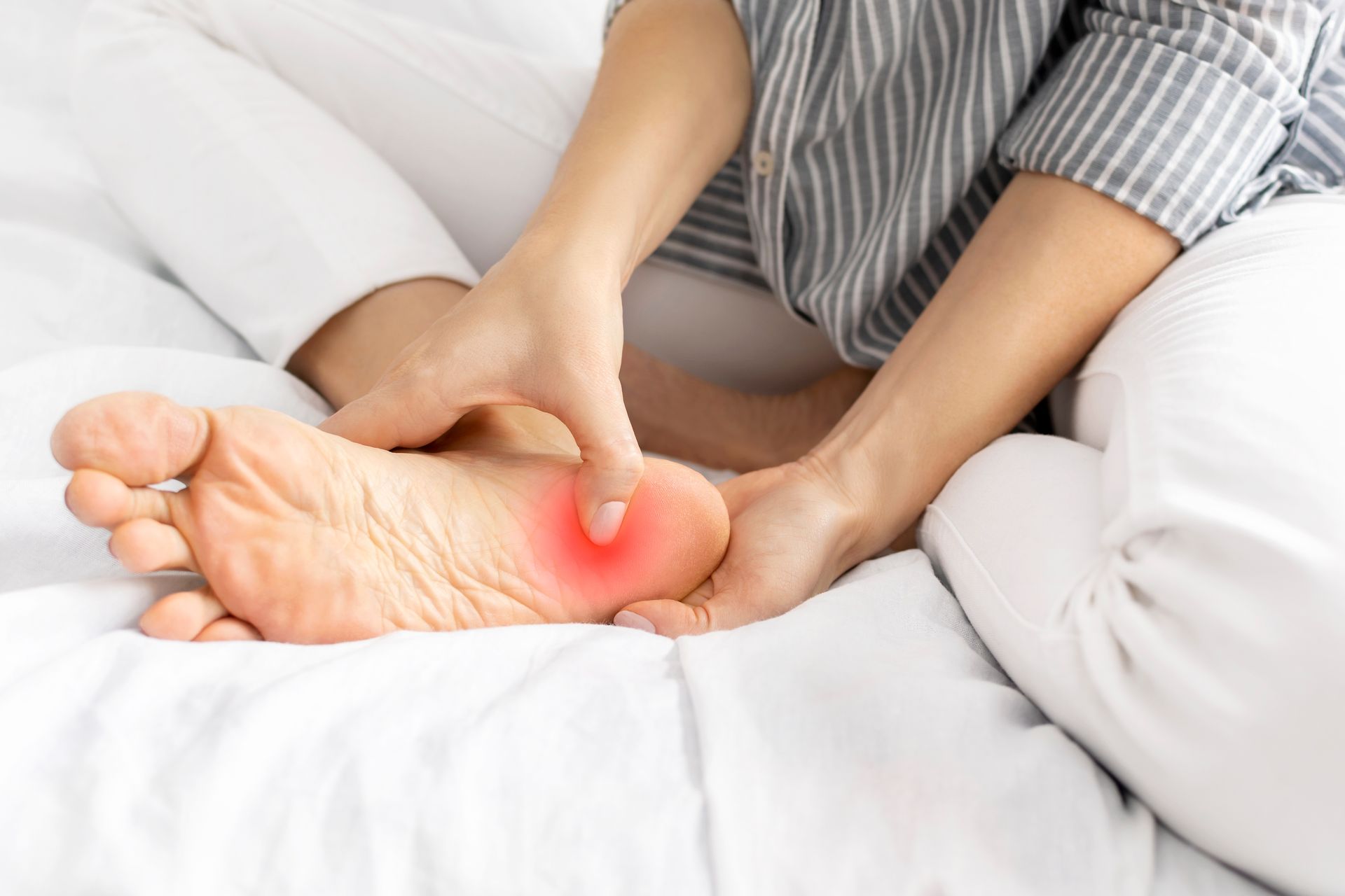 Person sitting on a bed, touching their heel with a red-highlighted area indicating pain.