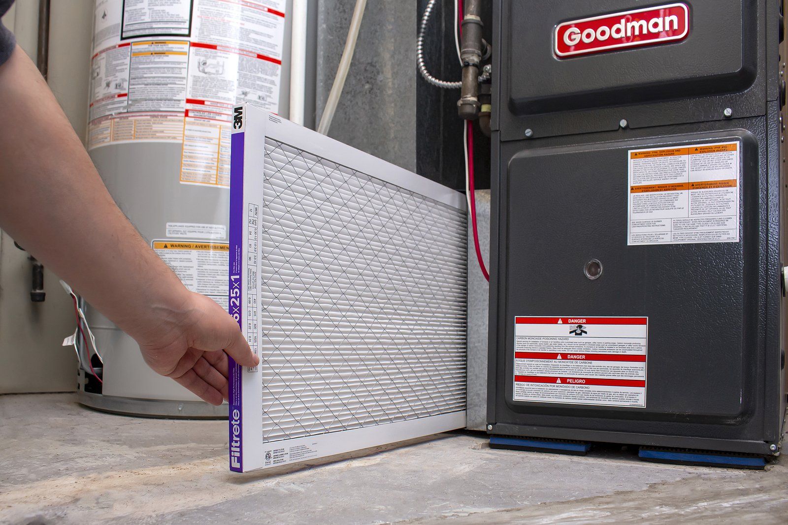 Hand replacing a furnace air filter beside a Goodman HVAC unit in a utility room