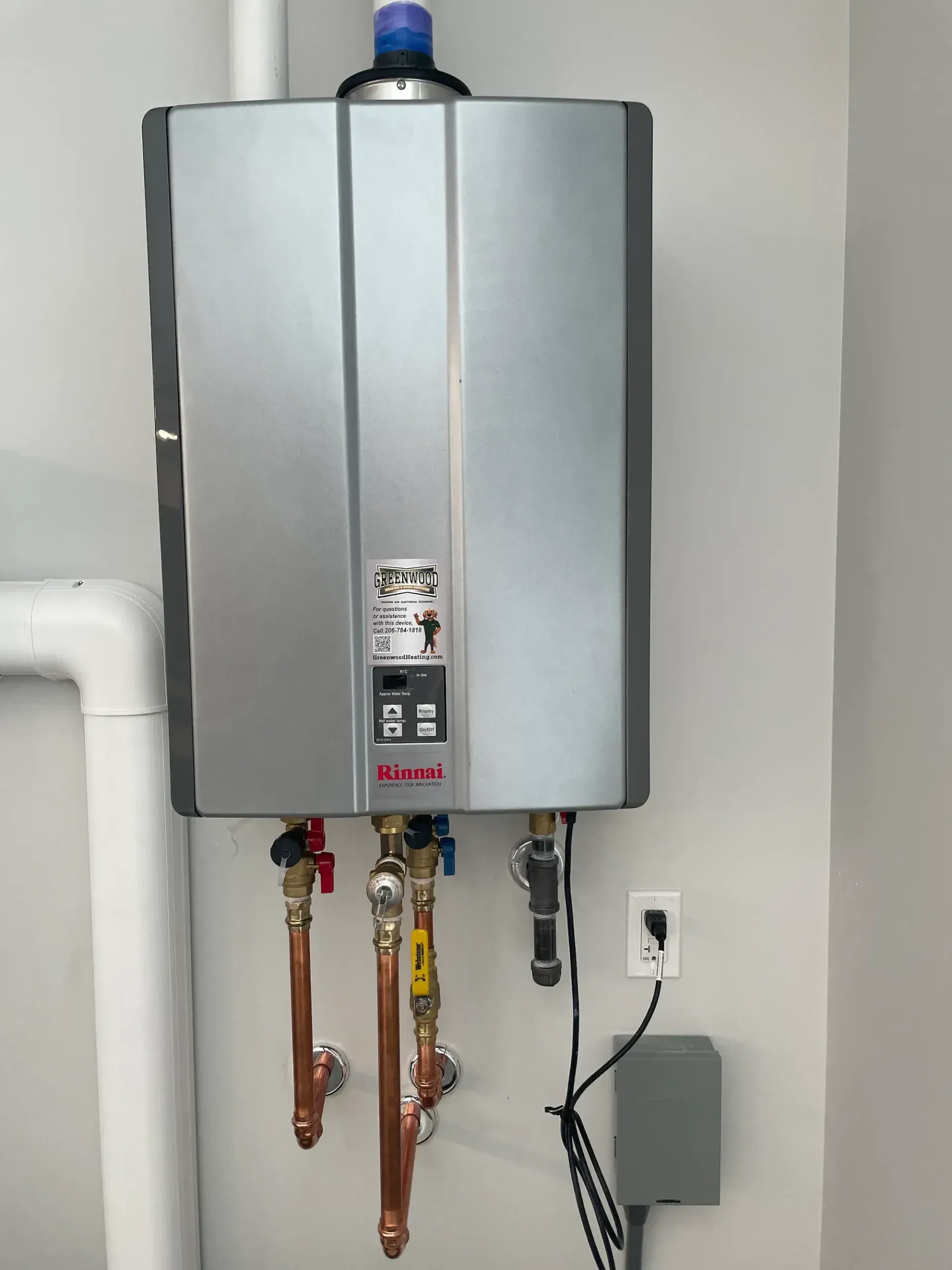 Wall-mounted silver water heater with pipes and electrical hookup in a utility room