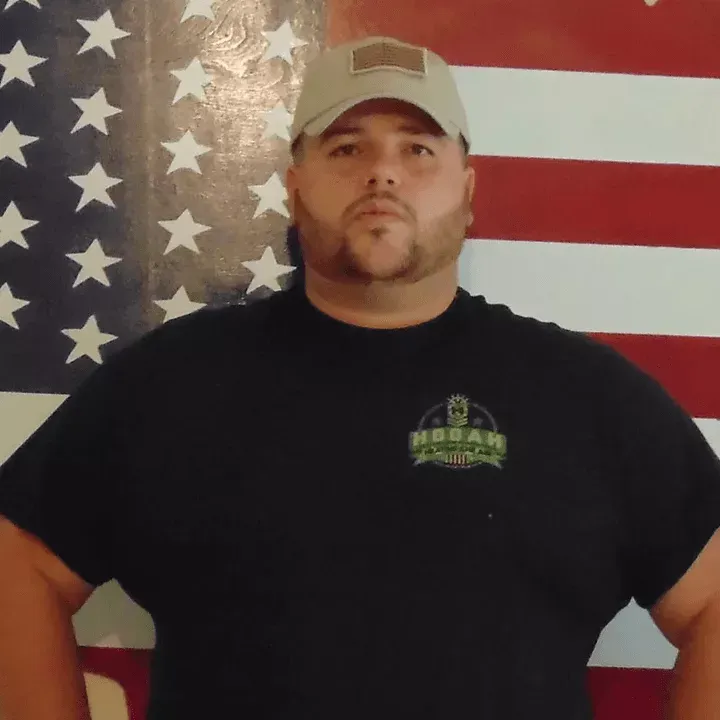 Sergio Goody Man standing before a large American flag, wearing a beige cap and black T-shirt.