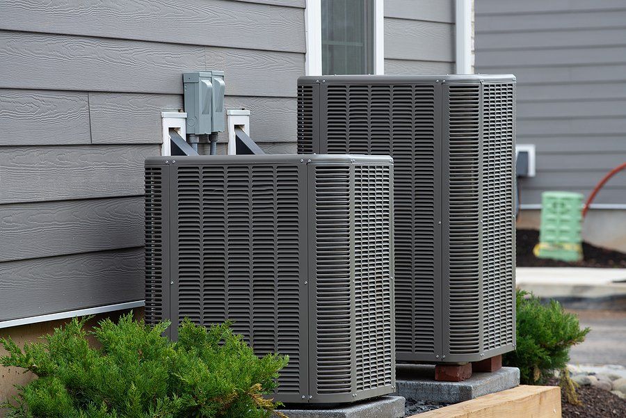 Two gray outdoor HVAC units beside a house, with green shrubs in front.