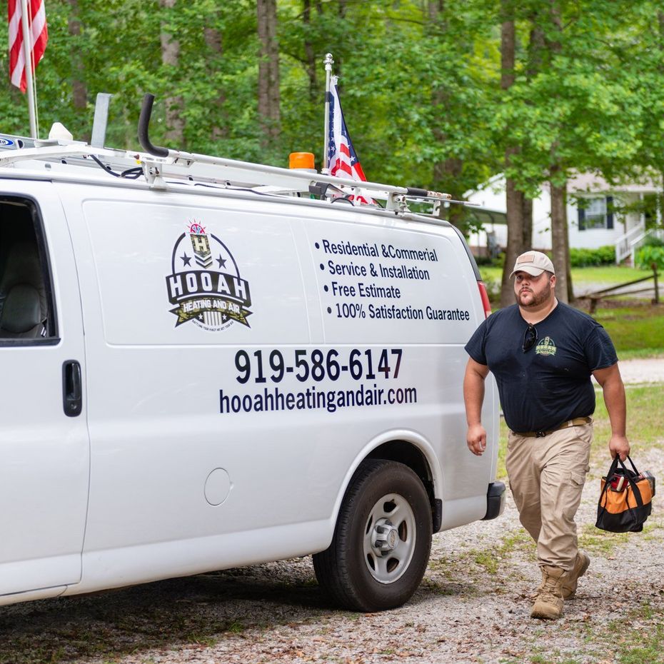 HVAC Services in Holly Springs, NC White service van with logo and phone number; man walks beside it carrying tools in a wooded driveway.