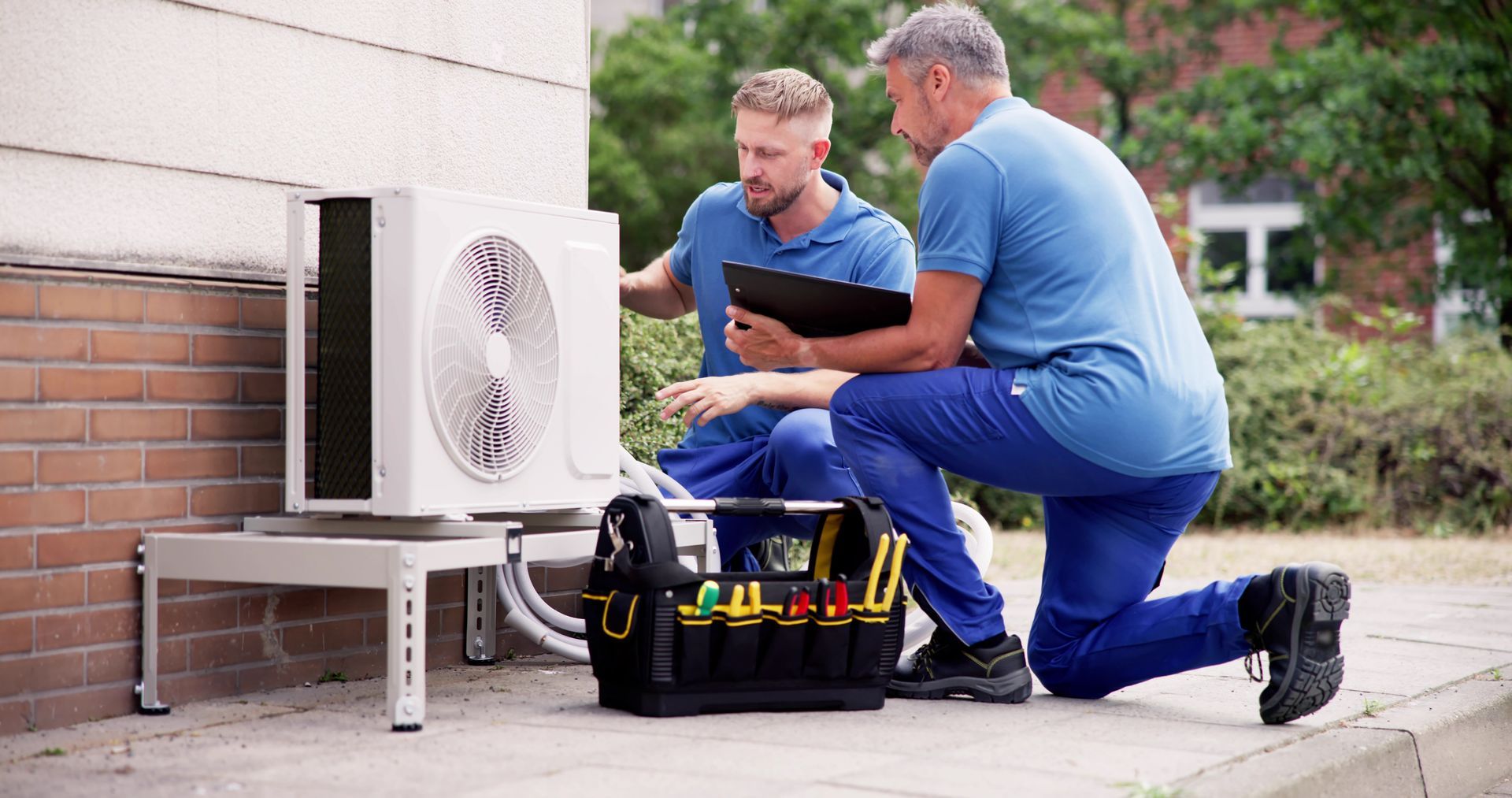 New Furnace Installations in Garner, NC Two technicians kneel beside an outdoor AC unit, checking it with a tool bag on the patio.