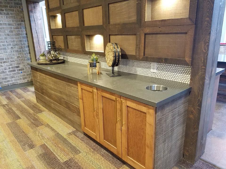 A wooden bar area with gray countertop, cabinets, and built-in shelving with decorative lighting.