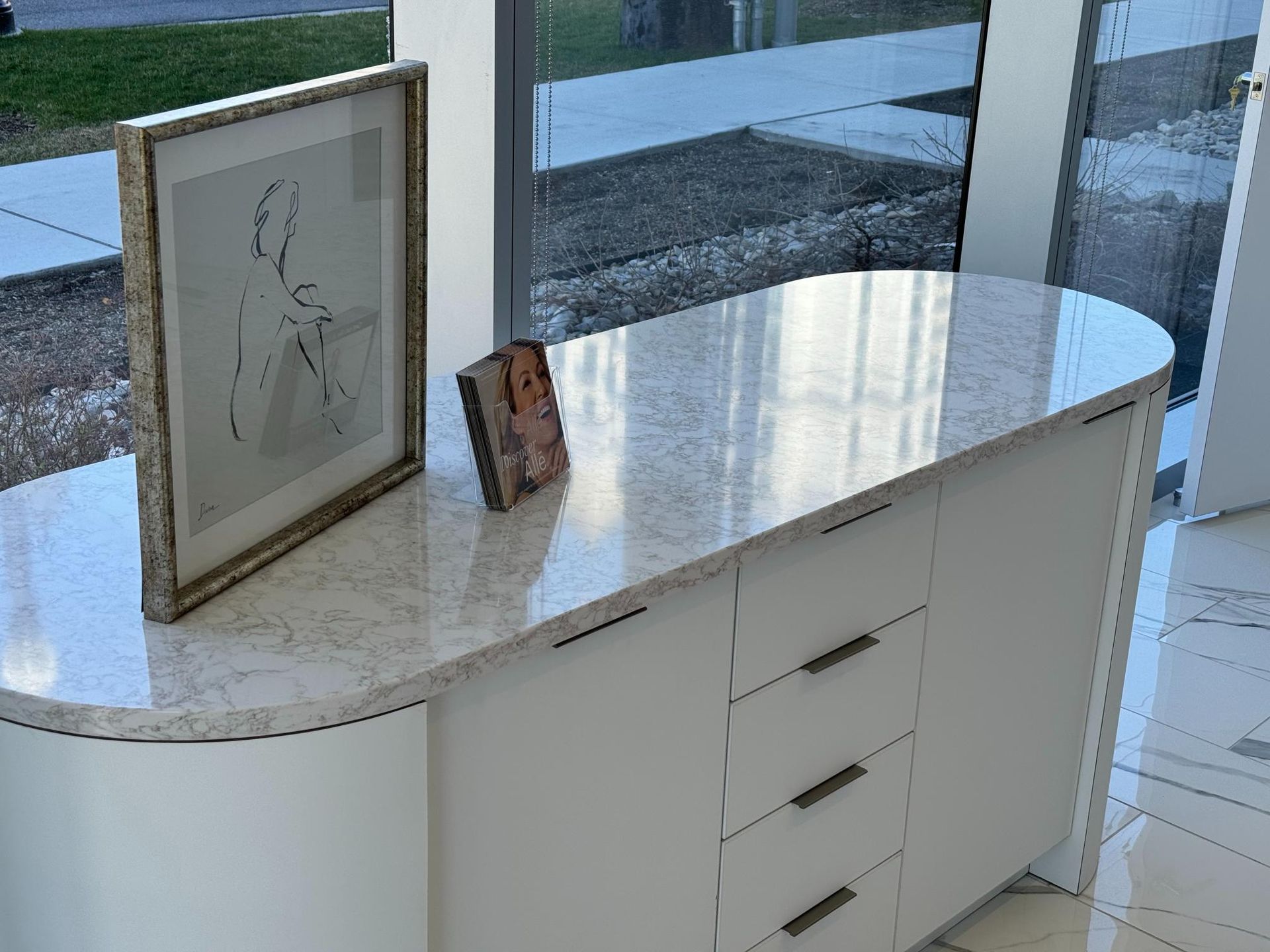 White cabinet with a marble top, artwork, and a small book sits by a large window.