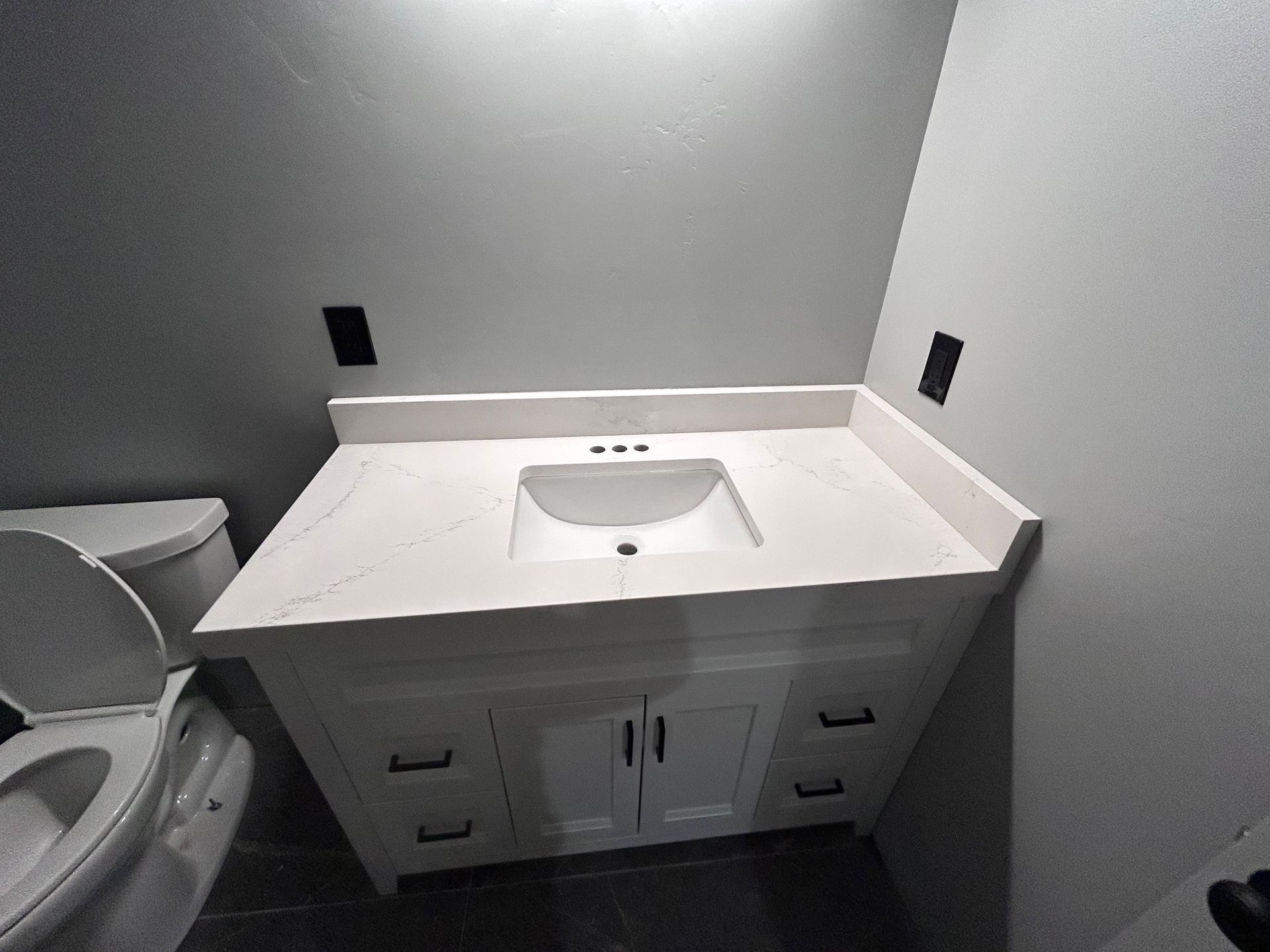 White bathroom vanity with sink and cabinets, next to a toilet. Gray walls.