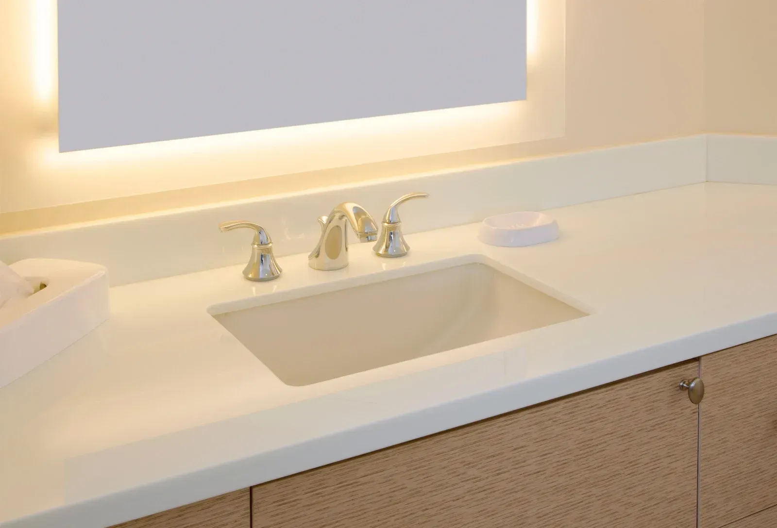 Bathroom sink with gold fixtures, under-lit mirror, and tan countertop, set on a light-colored wooden cabinet.