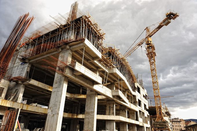 Un grande edificio in cemento in costruzione, con gru e impalcature, si staglia contro un cielo nuvoloso.