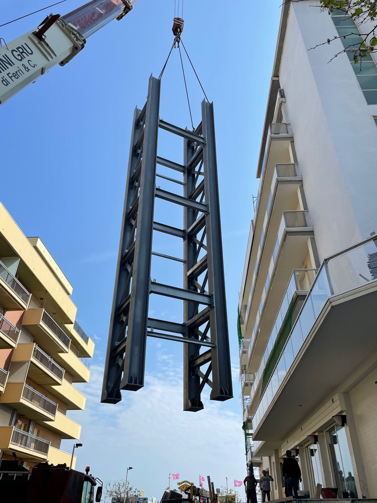 Una gru solleva un'alta struttura in acciaio tra gli edifici di una via cittadina, con gli operai visibili in cima.