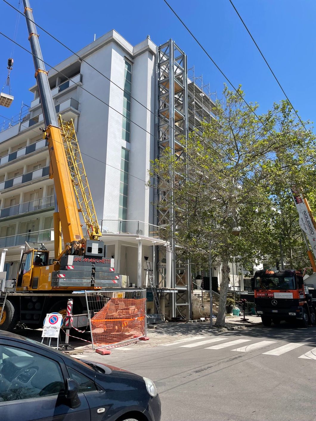Una gru solleva materiali accanto a un edificio in una via cittadina, con traffico e pedoni nelle vicinanze.