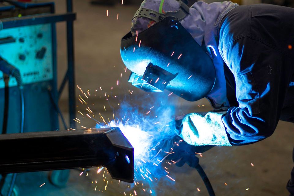 Un saldatore con indumenti protettivi sta saldando una trave di metallo, con scintille che volano in un'officina illuminata da una luce blu.