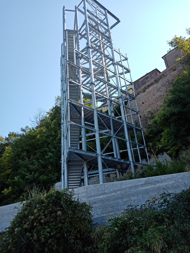 Un'alta torre di scale in metallo accanto a un muro di mattoni e alberi sotto un cielo azzurro.