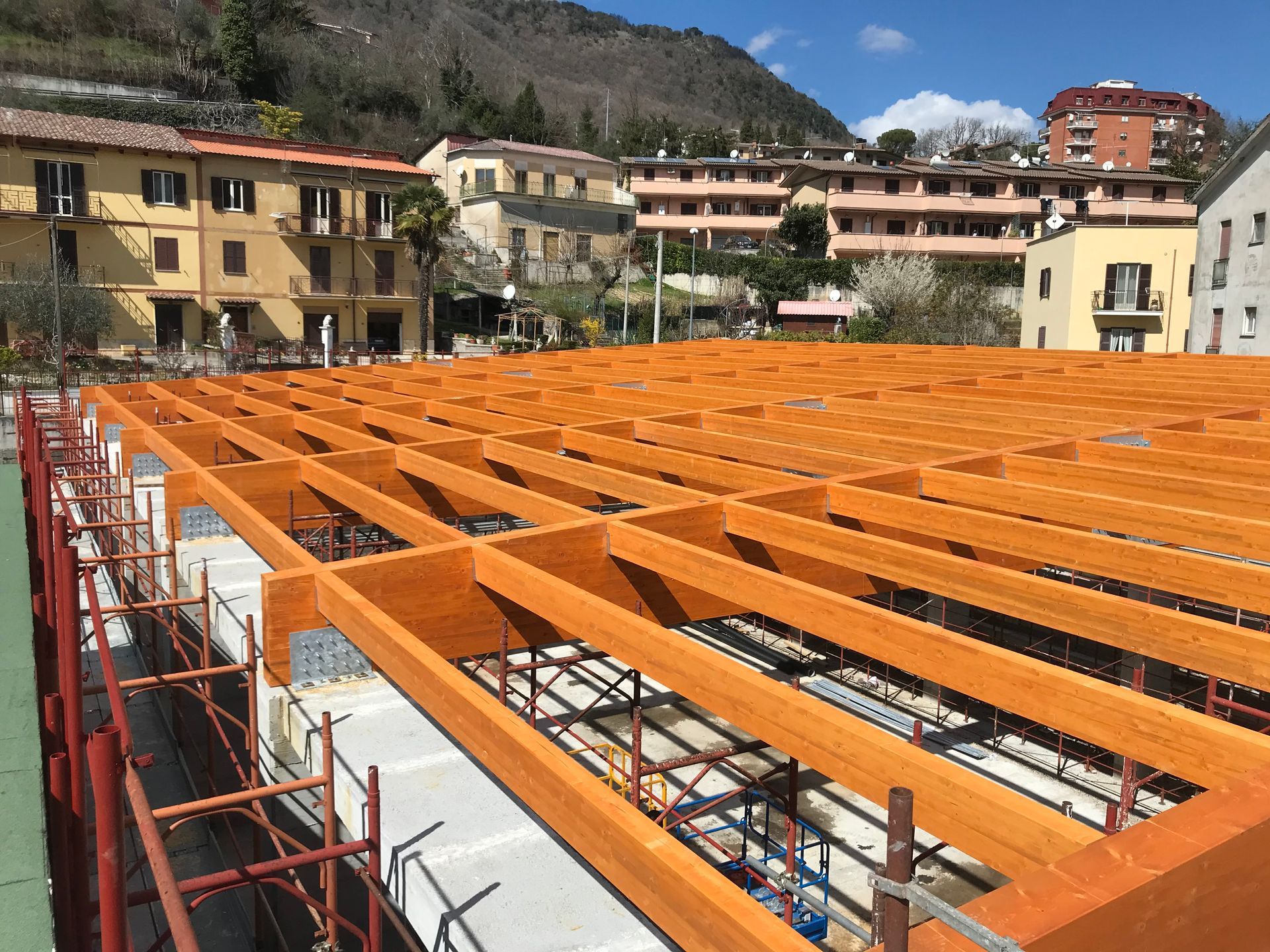 Cantiere edile con travi arancioni sul tetto di un edificio, circondato da impalcature e case arroccate sulla collina.