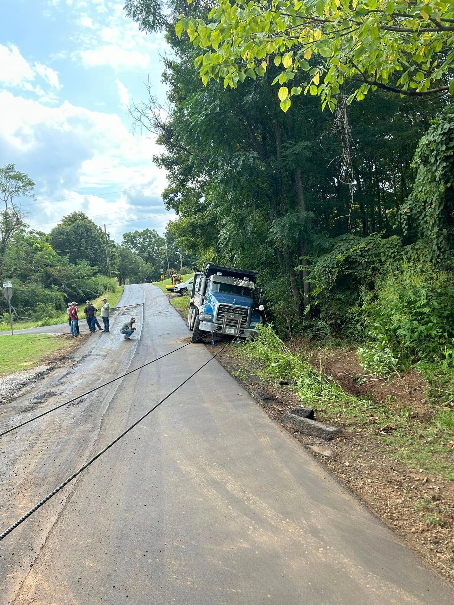 Damaged truck on a wet rural road with several people nearby and trees along the roadside