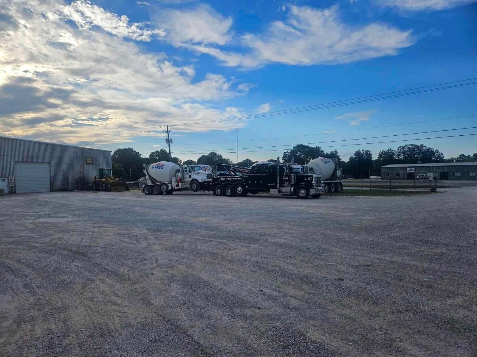 Concrete yard with several cement mixer trucks parked beside an industrial building under a cloudy blue sky