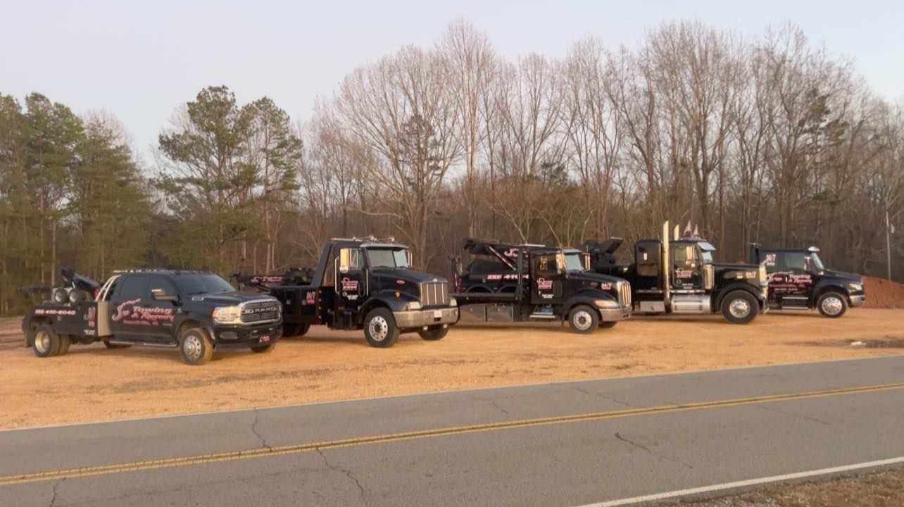 Several tow trucks and SUVs parked along a roadside near leafless trees and a highway