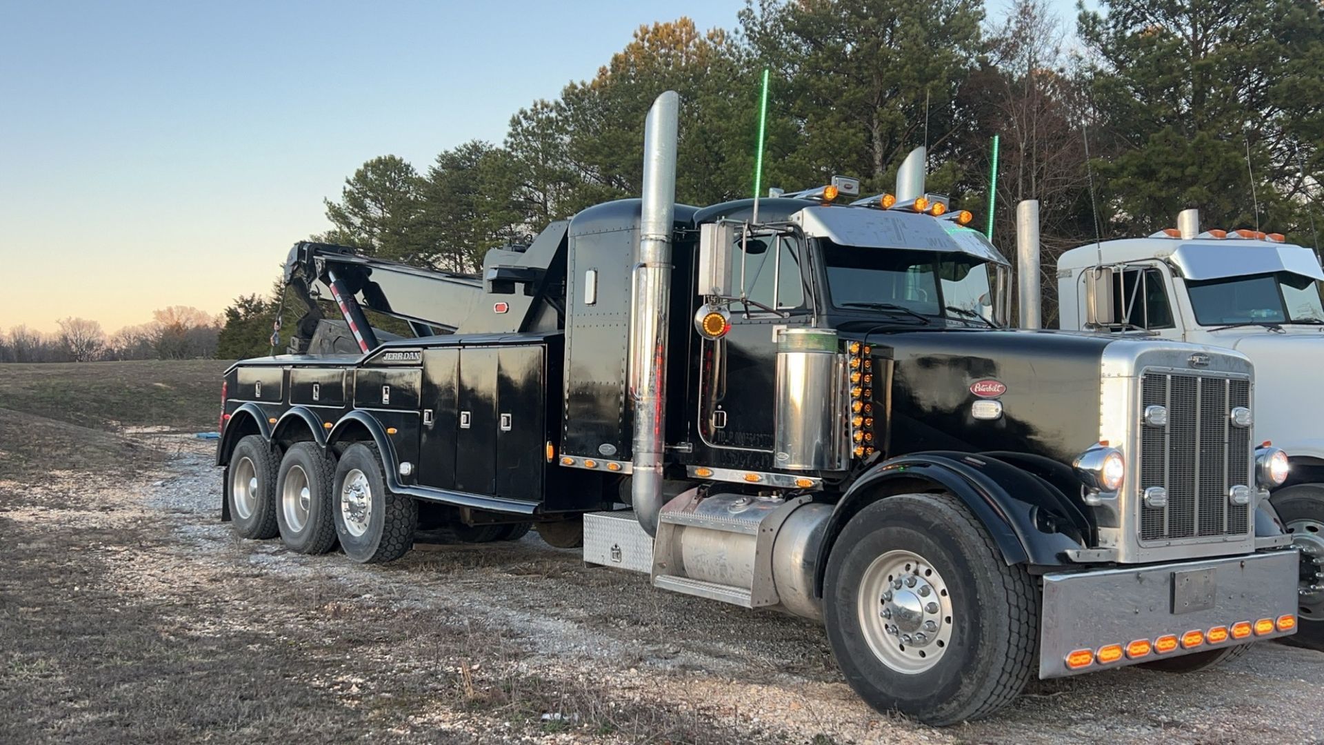 Black semi-truck with flatbed trailer parked on gravel at sunrise near trees.