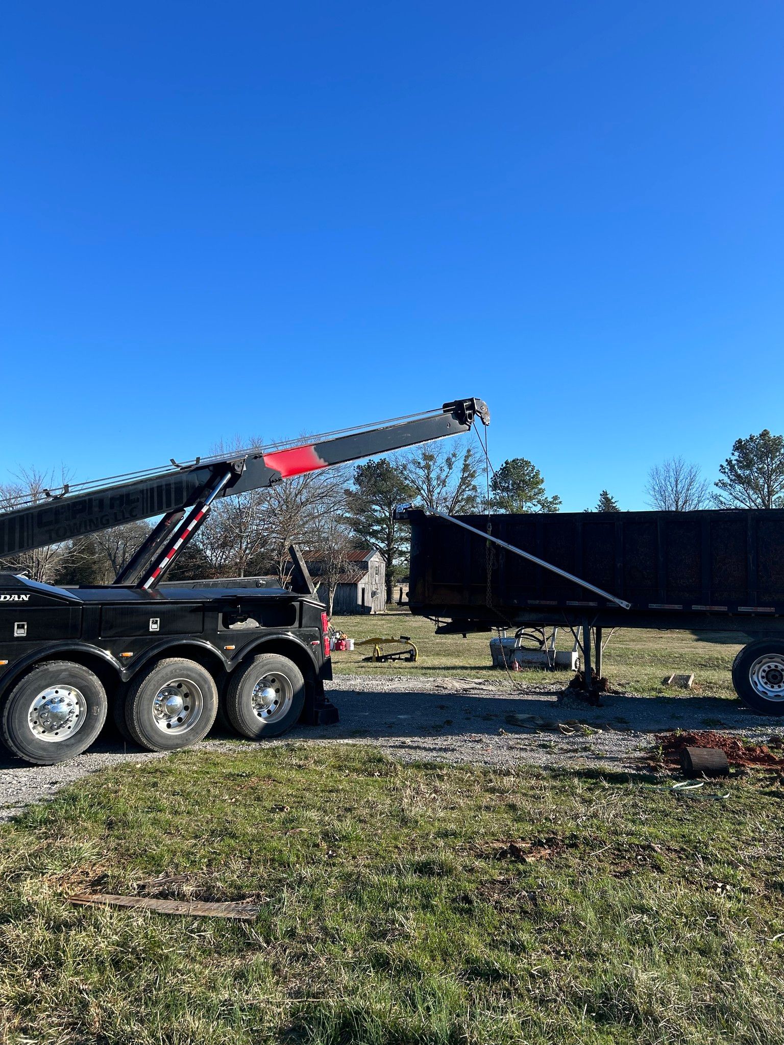 Tow truck lifting a black trailer in a grassy yard under a clear blue sky