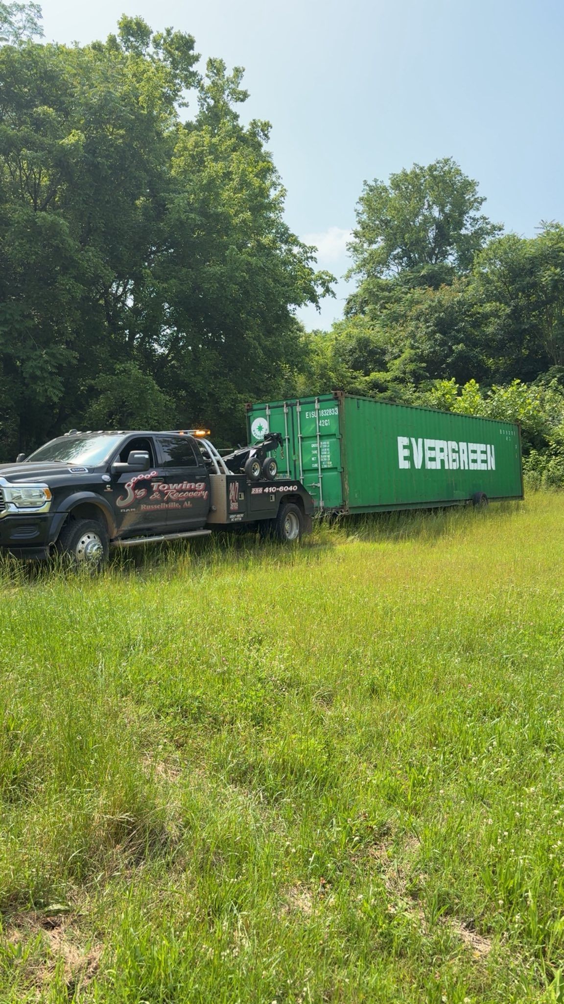 Black pickup truck towing a green dumpster on grassy roadside with trees in the background