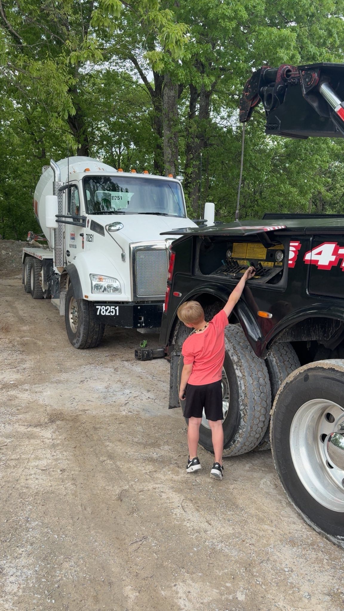 Child in red shirt standing beside a black truck and white semi-truck on a gravel road under trees