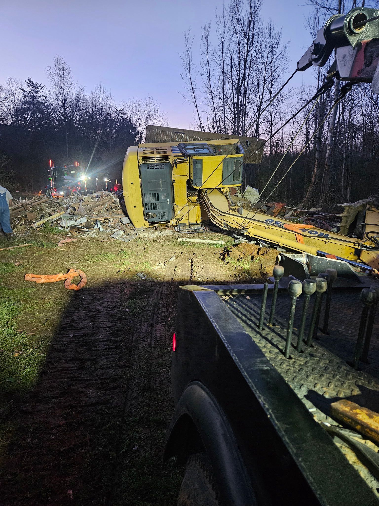 Overturned yellow vehicle on a roadside at dusk, with emergency lights and debris nearby
