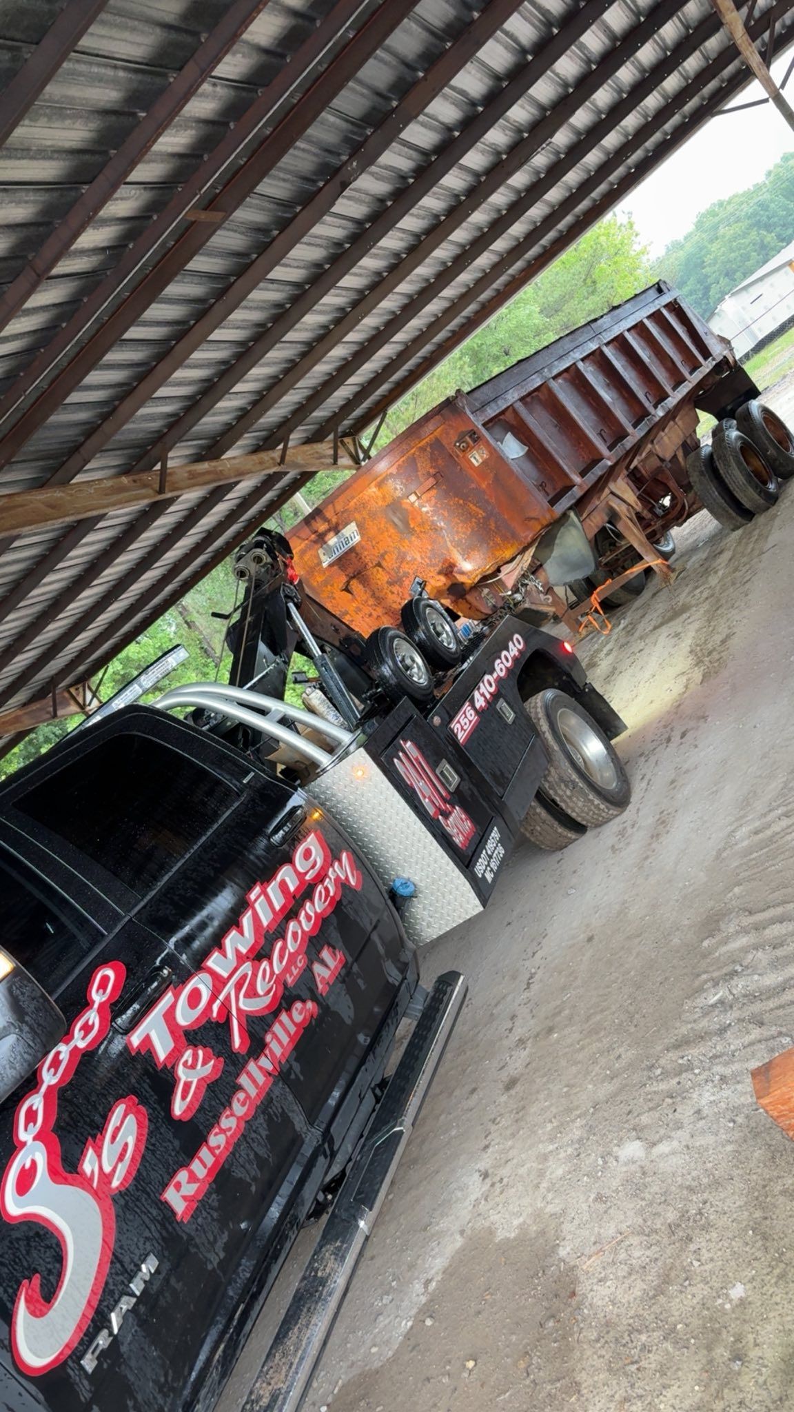 Covered parking area with a black truck and orange dump truck parked side by side on gravel.