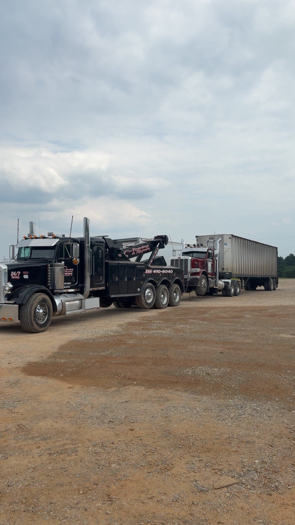 Black semi truck hauling a long flatbed trailer parked on a gravel lot under a cloudy sky