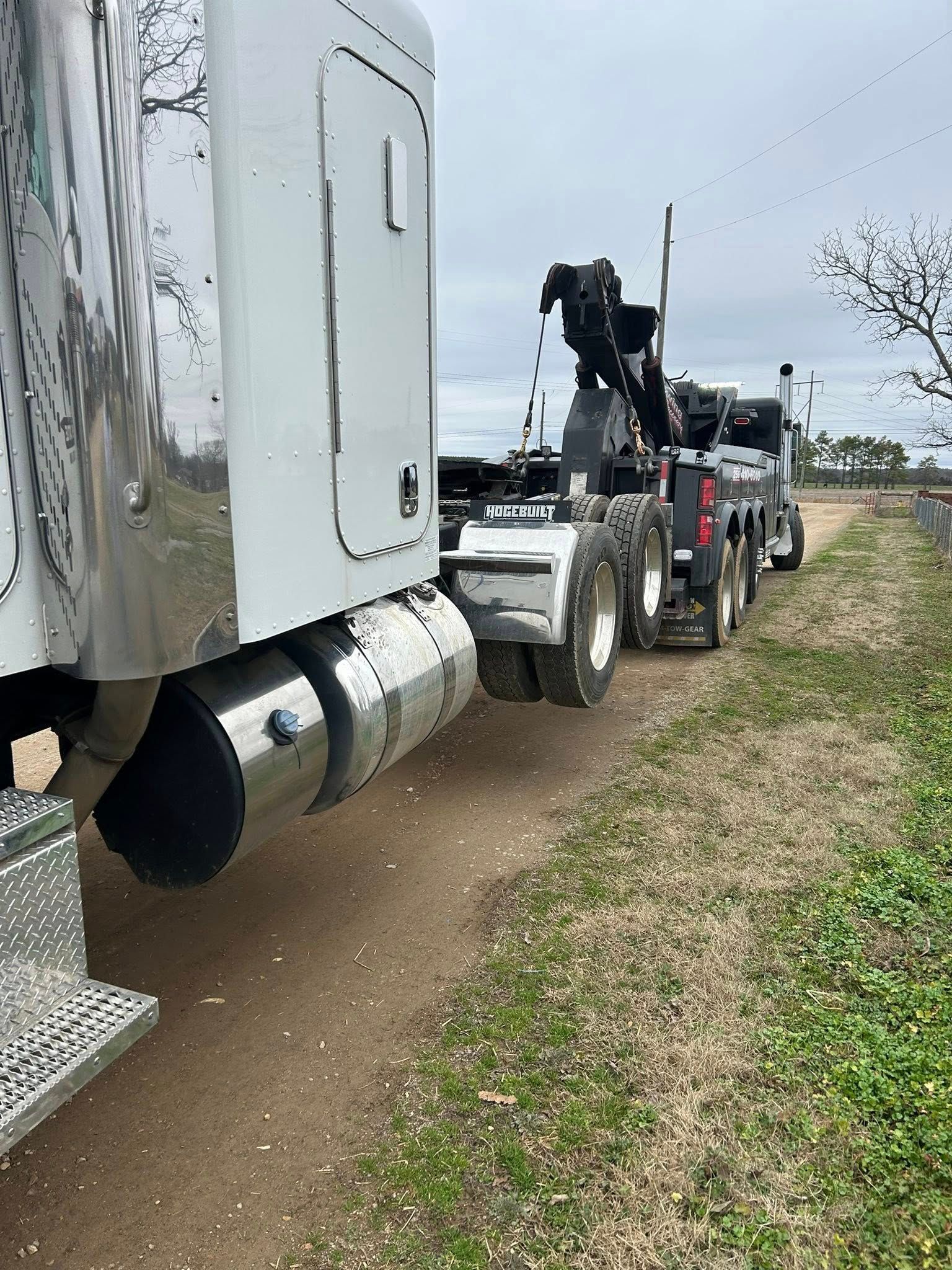 Silver semi-truck with a crane attachment parked on a dirt shoulder beside a road.