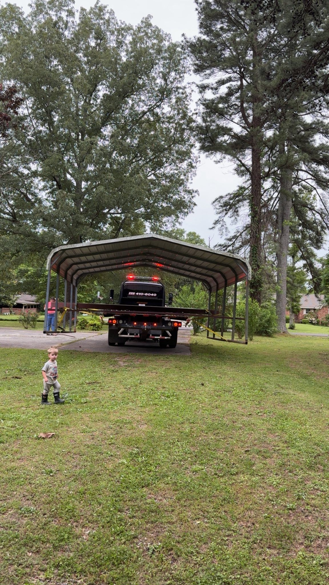 Black pickup truck under a carport in a grassy yard, with two children nearby and tall trees around.