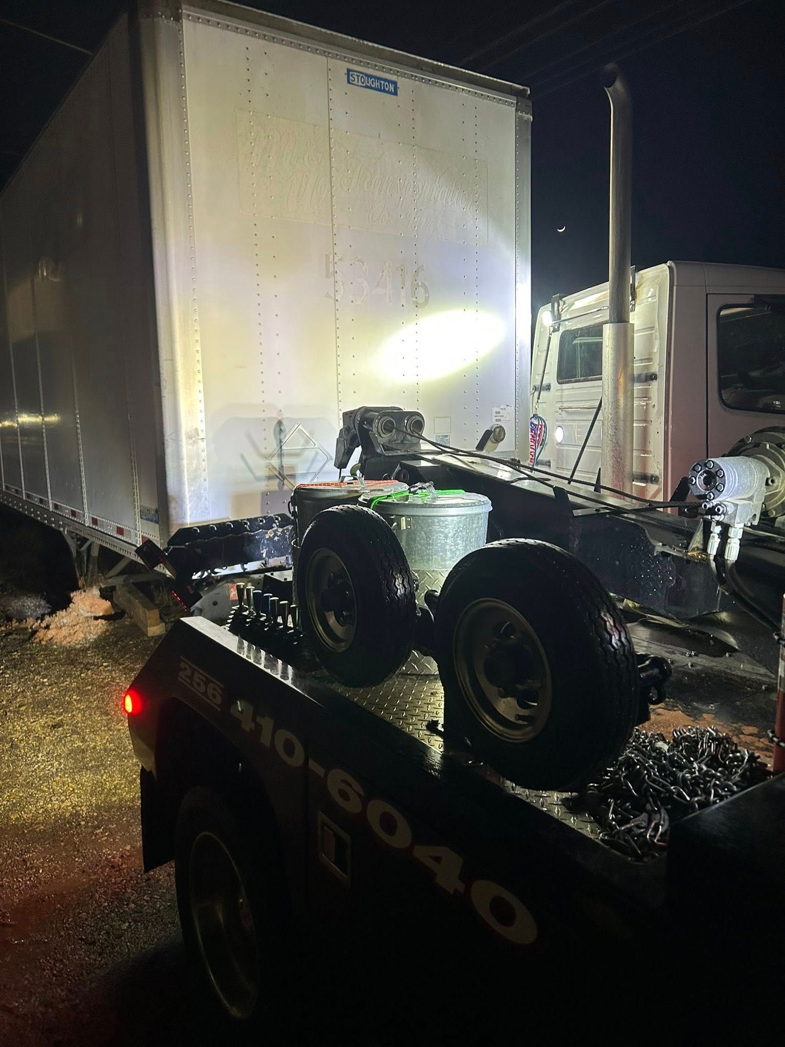 Tow truck lifting an overturned car at night beside a large box truck in the rain