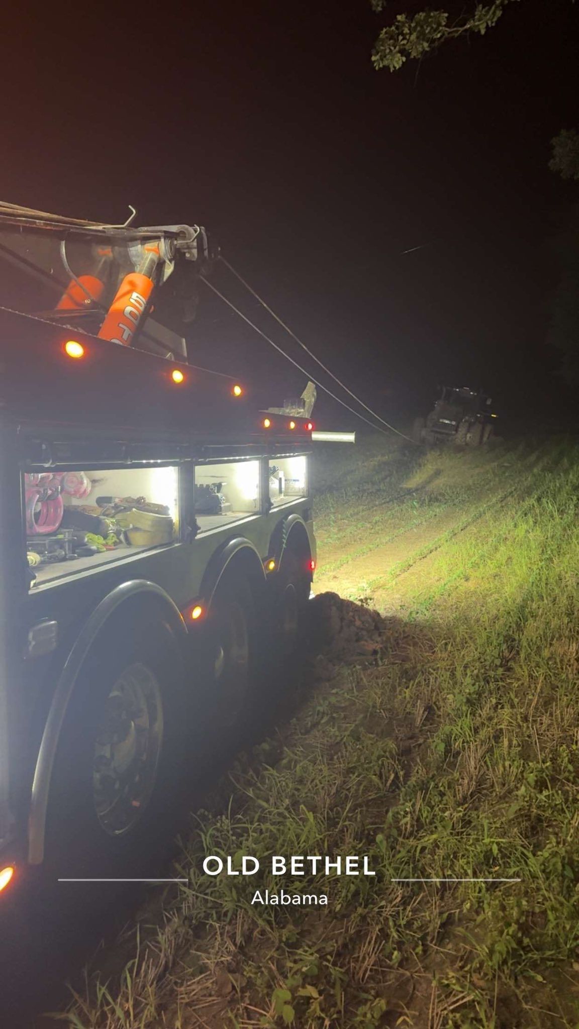 Tow truck with flashing lights at night on a grassy roadside near a wooded area.