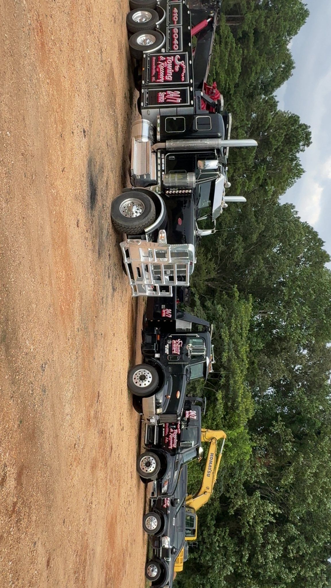 Several tow trucks and heavy-duty trucks parked in a gravel lot by trees