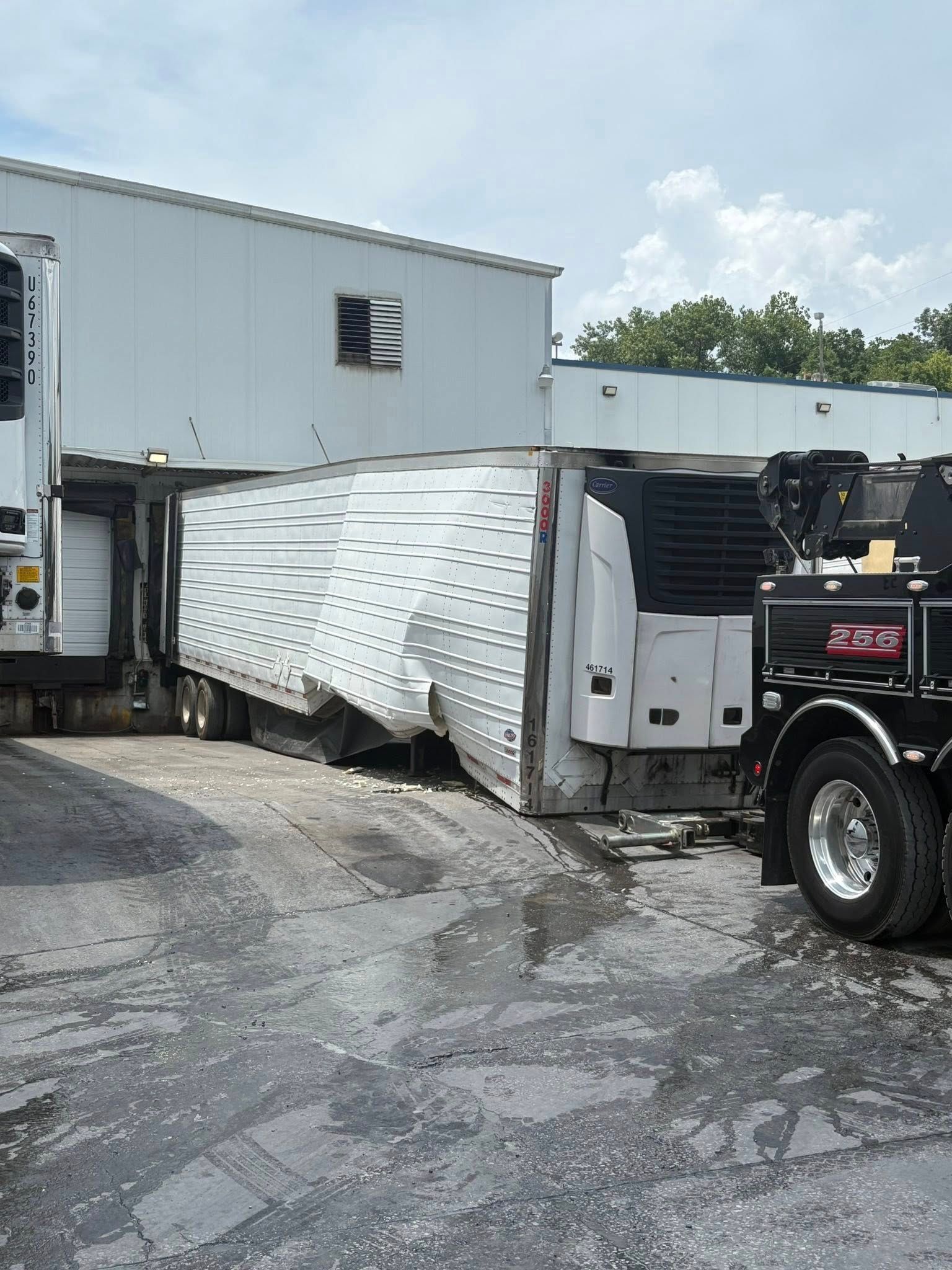 Crushed white box truck wedged between trailers in a damaged lot.