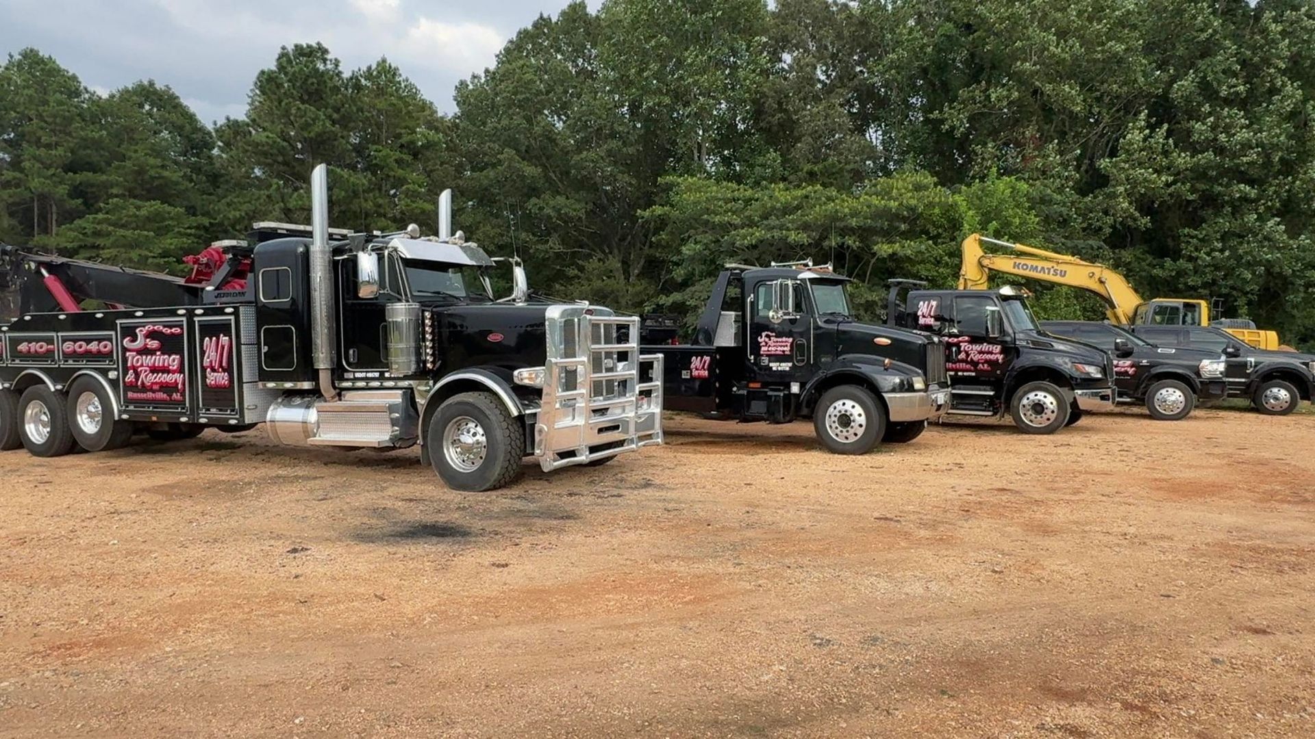 Line of black tow trucks parked on a dirt lot beside trees, with one yellow excavator in back