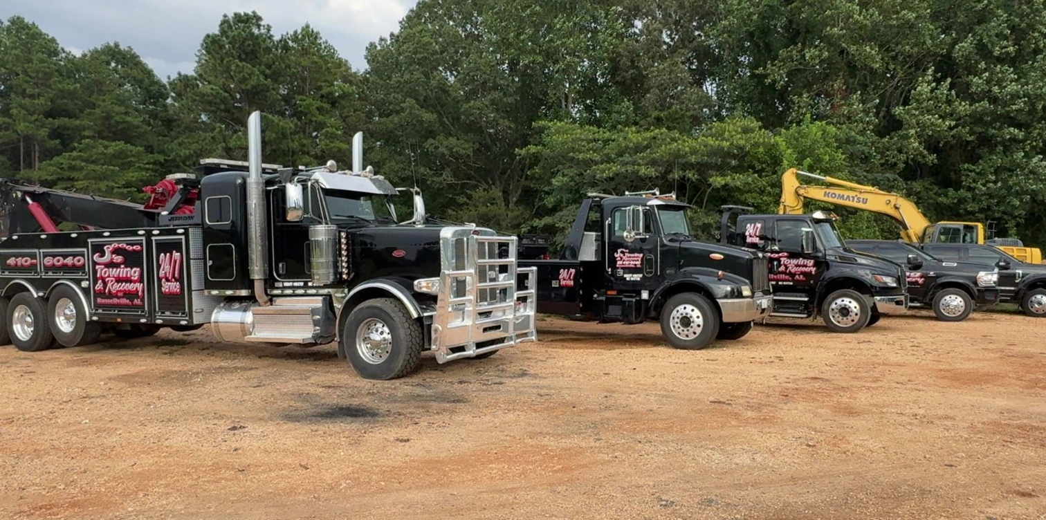 Row of black tow trucks and equipment trucks parked on gravel under trees