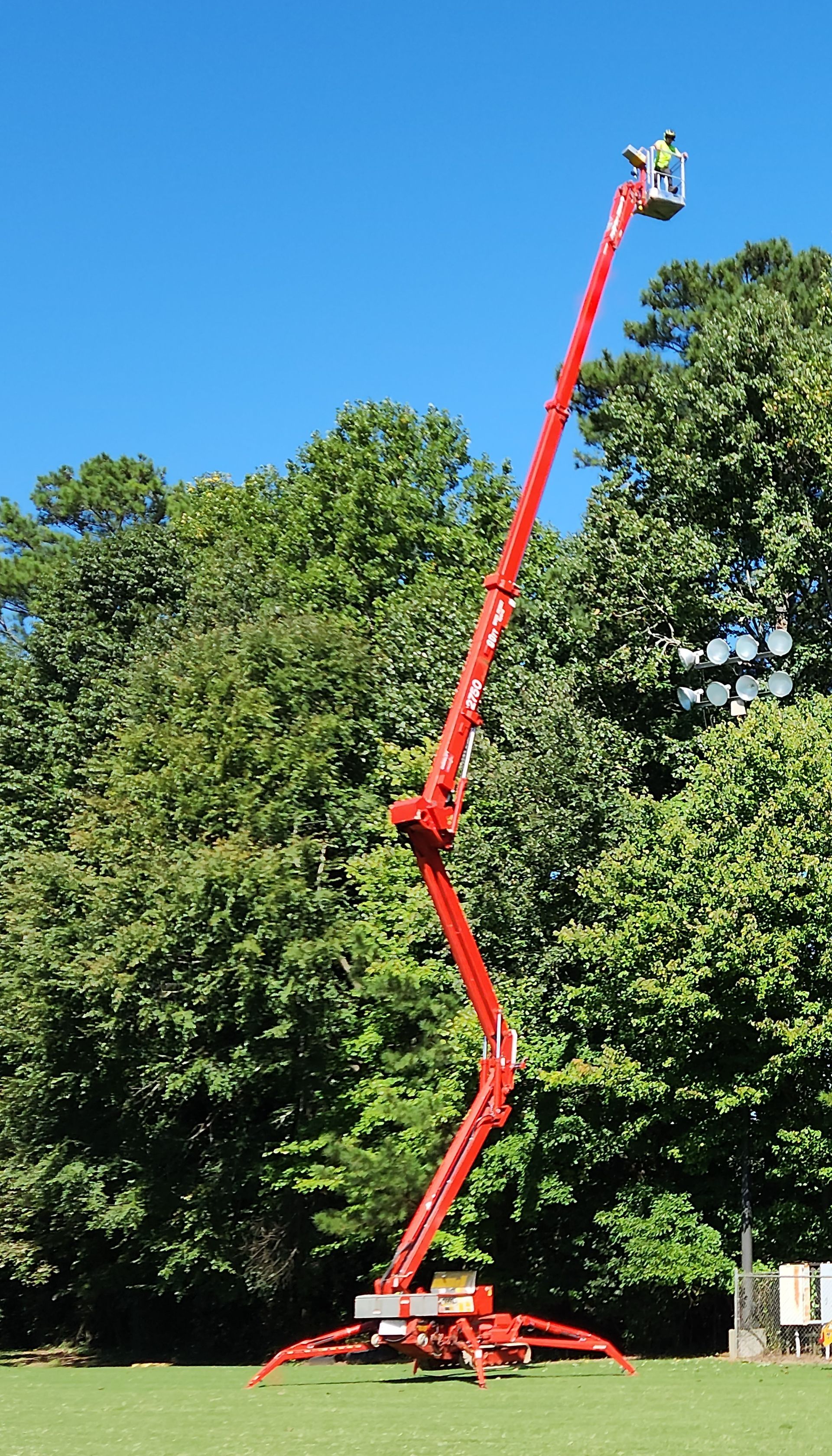 A person in a harness stands in the basket of a red articulating boom lift extended high into the air in a park.