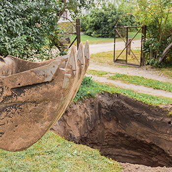 A large, rusty excavator bucket hovering over a deep, freshly dug rectangular trench near a dirt path and metal gate.