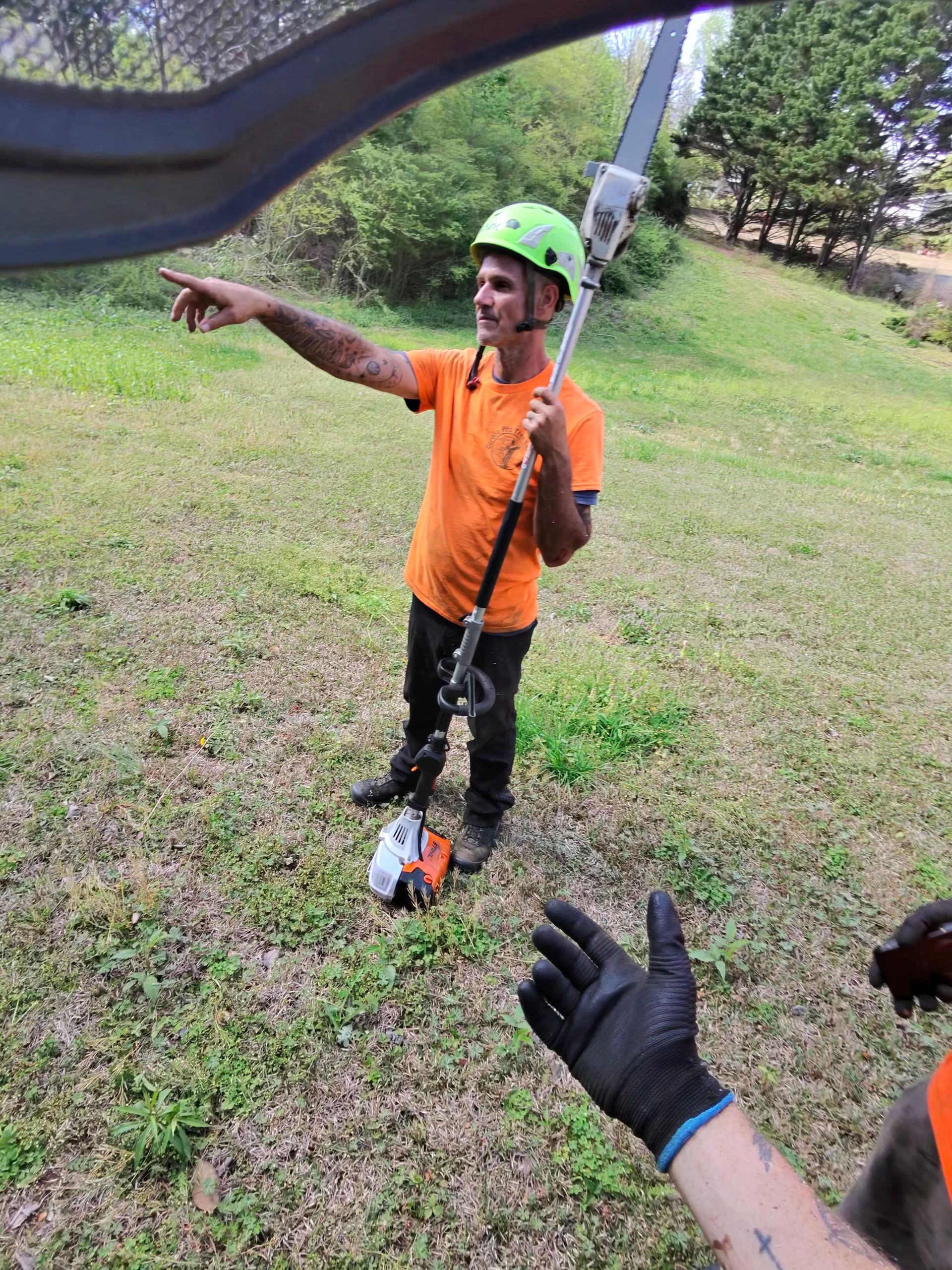 A person in a bright orange shirt and neon green hard hat holds a pole saw while pointing toward a field.
