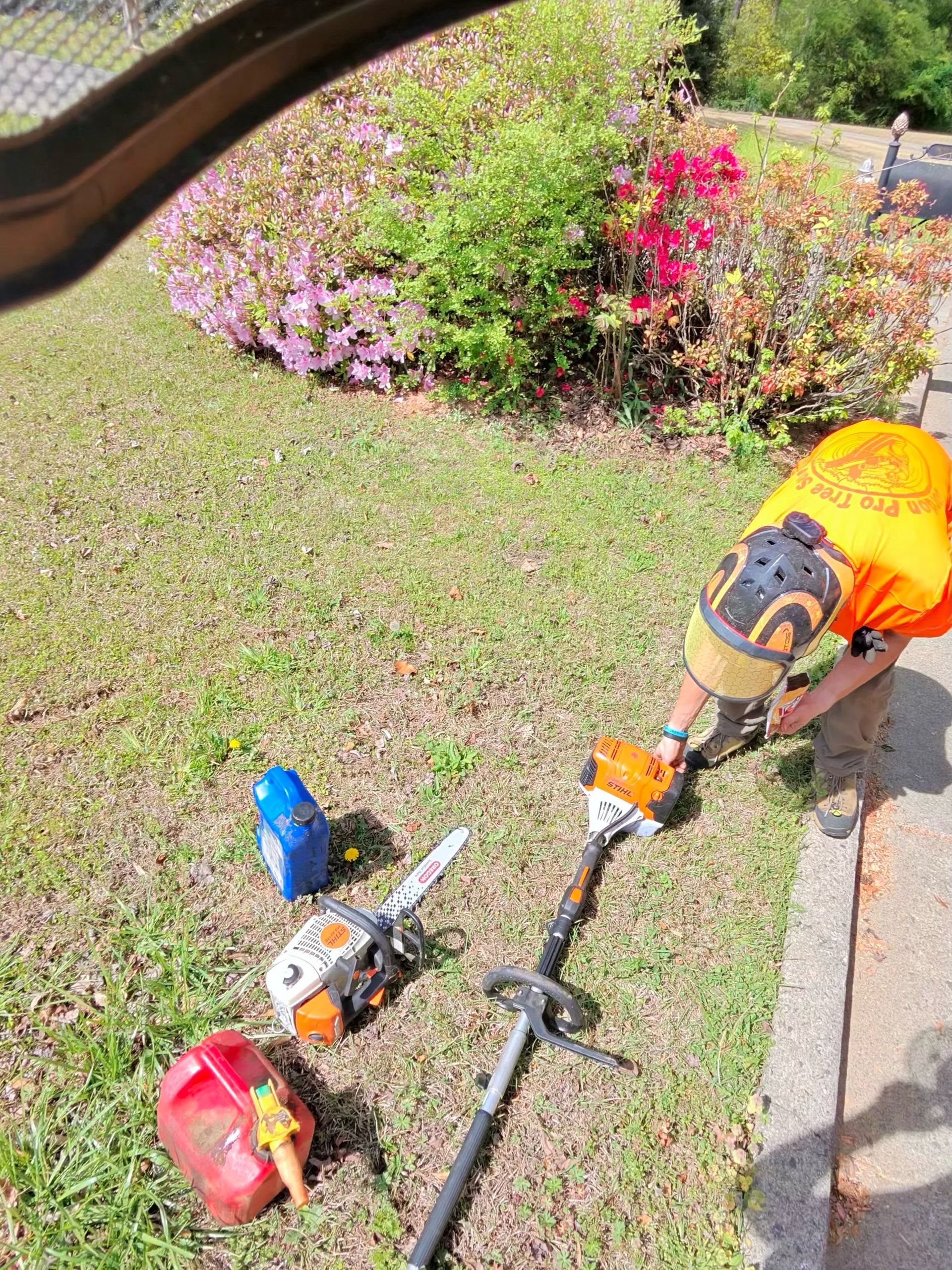 A person wearing a helmet and orange shirt works on a long-reach pole hedge trimmer beside a chainsaw and fuel containers.