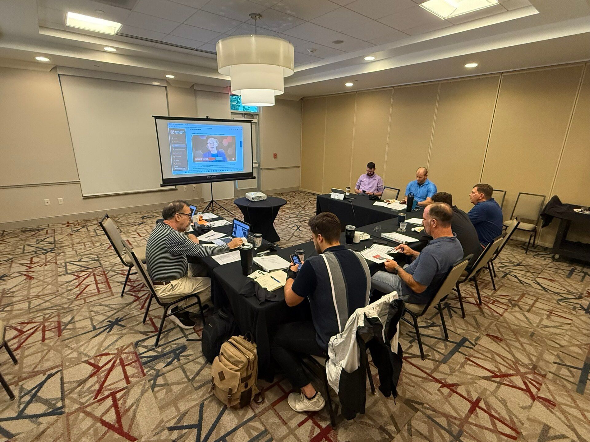 A meeting in a conference room with people seated around a table, a projector screen displays a weather map.