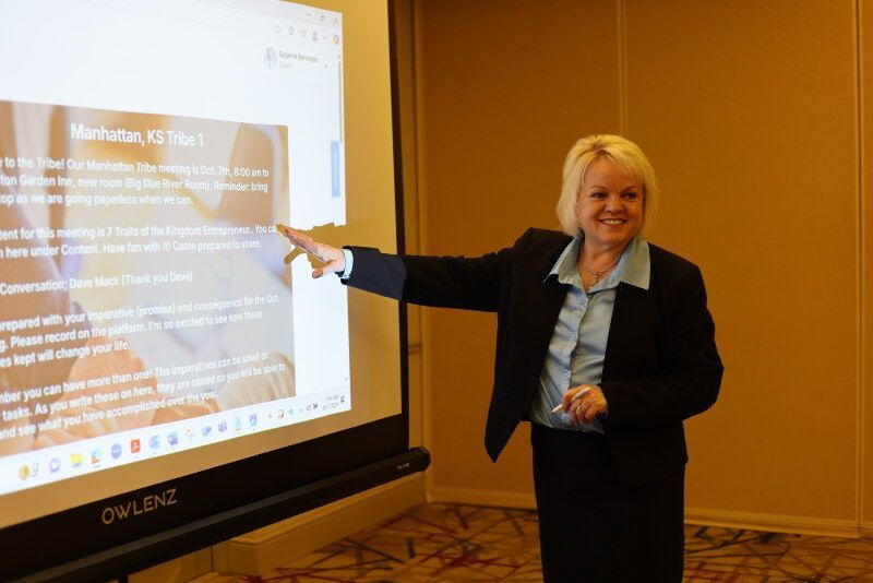 Woman pointing at a screen in a presentation. She's smiling, wearing a suit in a conference room.