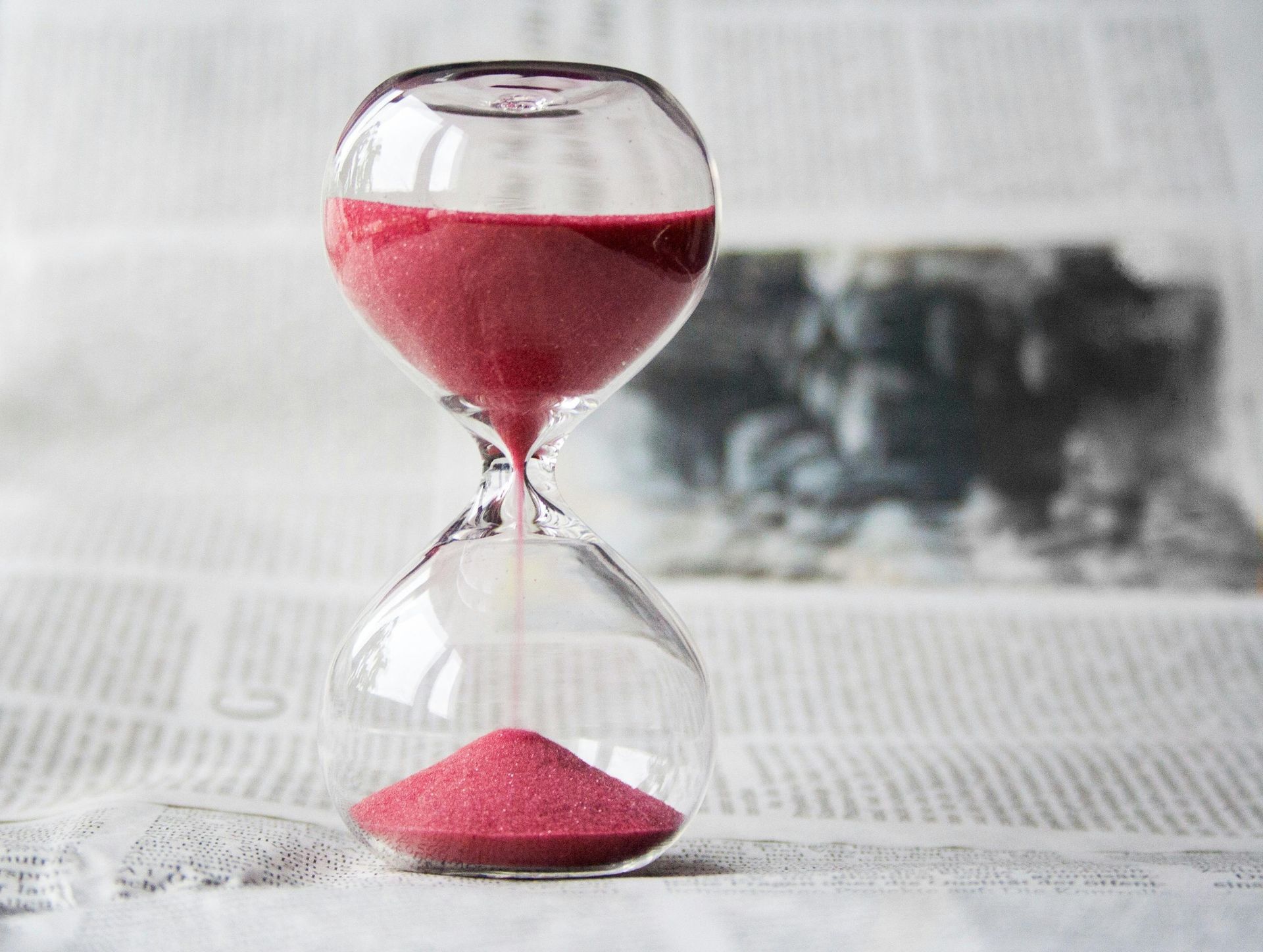Hourglass with pink sand, sand flowing from top to bottom, resting on newspaper.