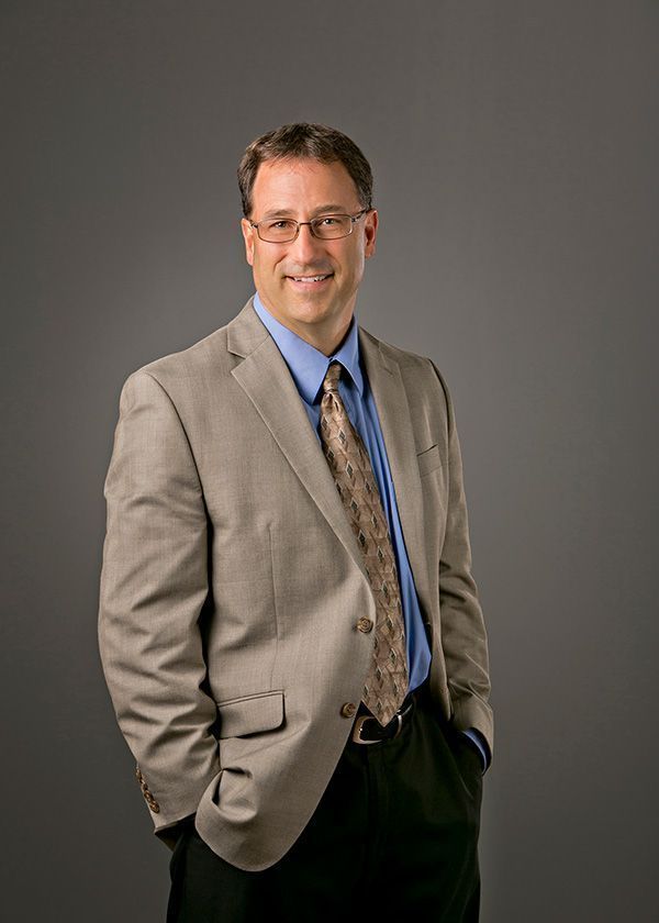 Man in a blazer and glasses, smiling, hands in pockets. Gray background.