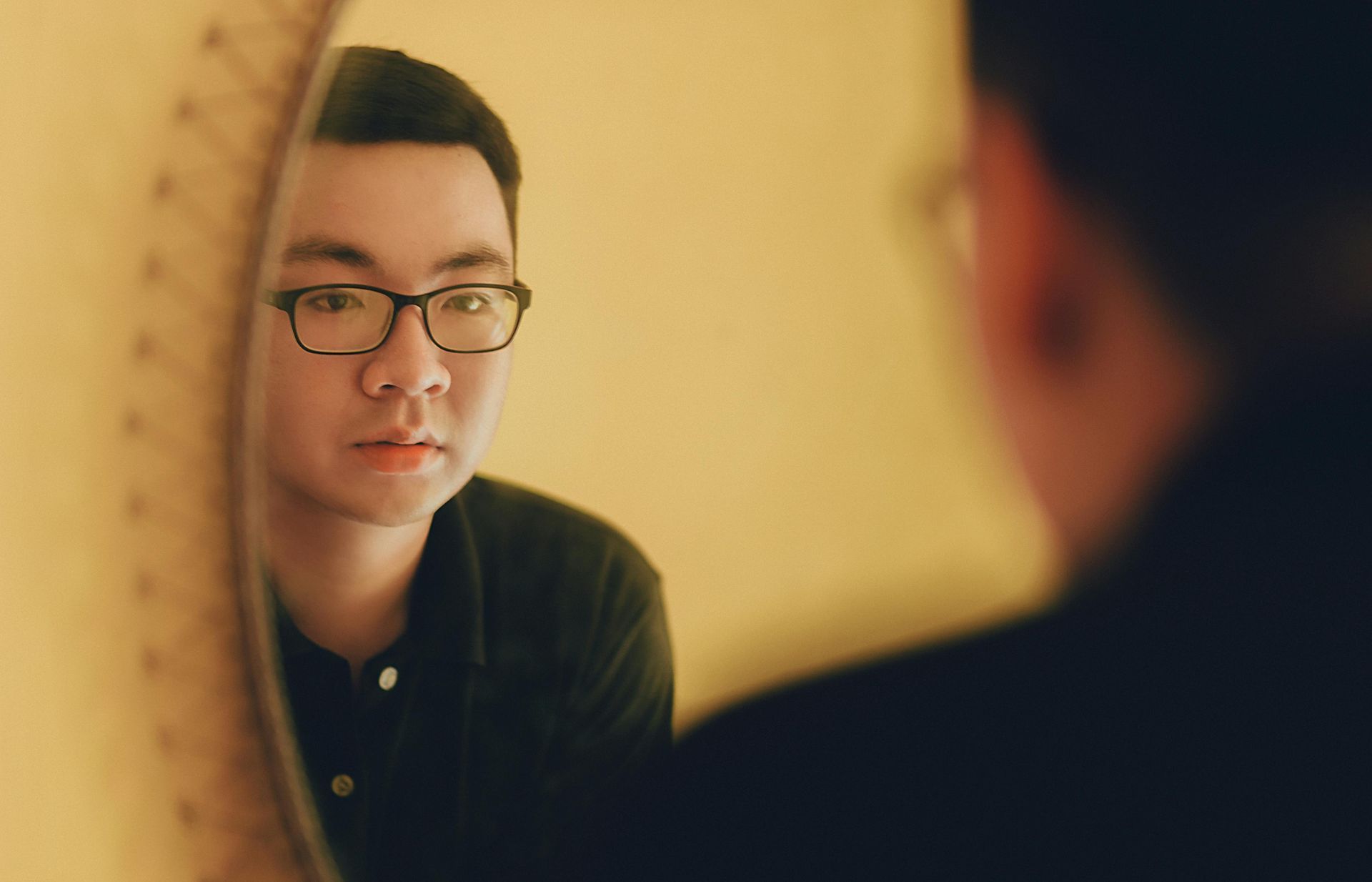 Man in glasses looking in a round mirror, yellow wall in the background.