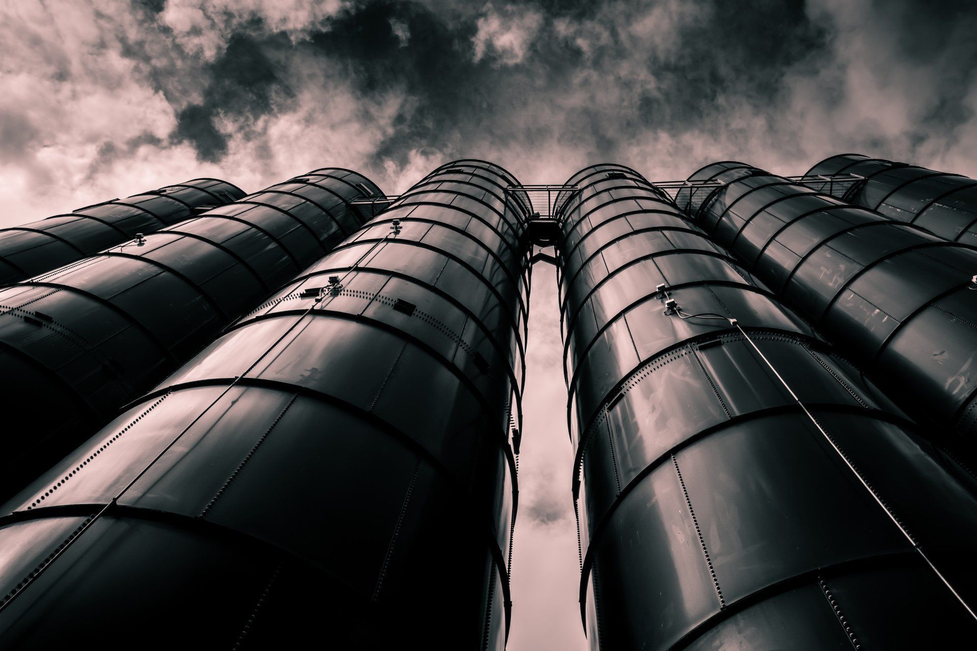 Dark, low-angle shot of tall metal industrial silos against a cloudy sky.