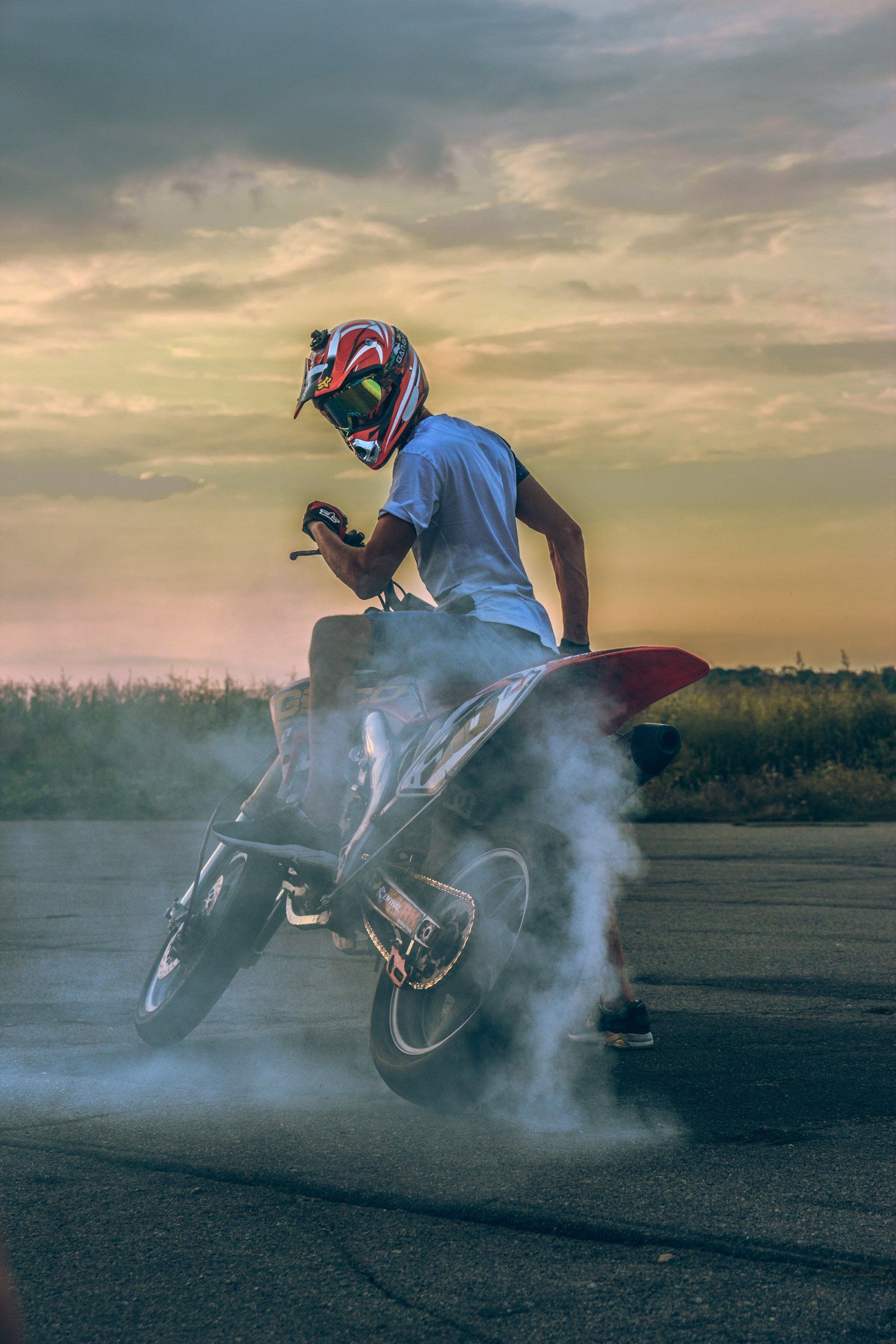 Motorcyclist doing a burnout, kicking up smoke against a sunset sky.