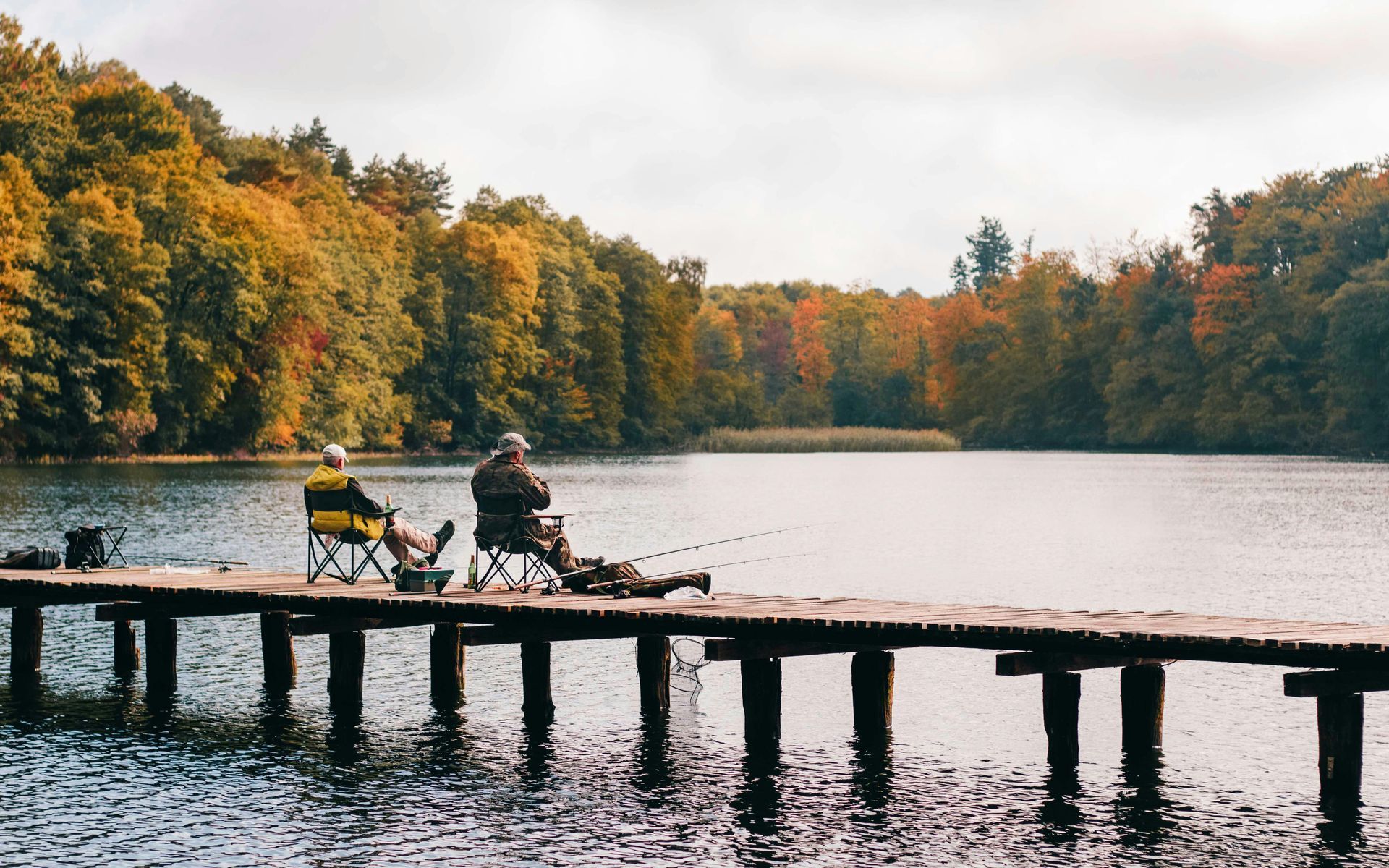 Two people fishing from a wooden pier on a calm lake surrounded by trees with autumn foliage.