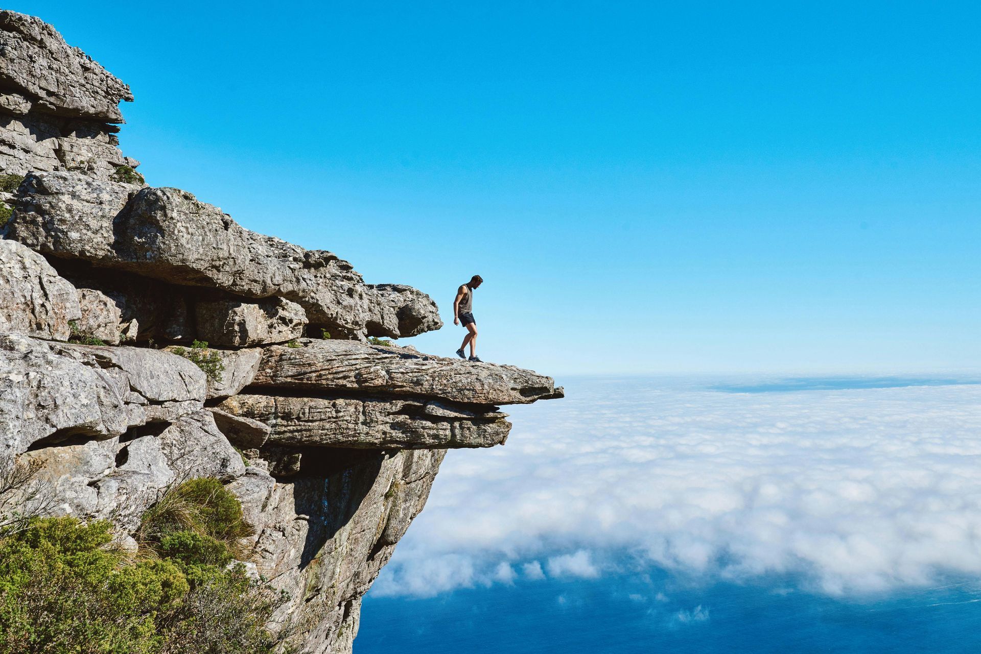 Person stands at edge of cliff, looking out at cloudy horizon on a sunny day.